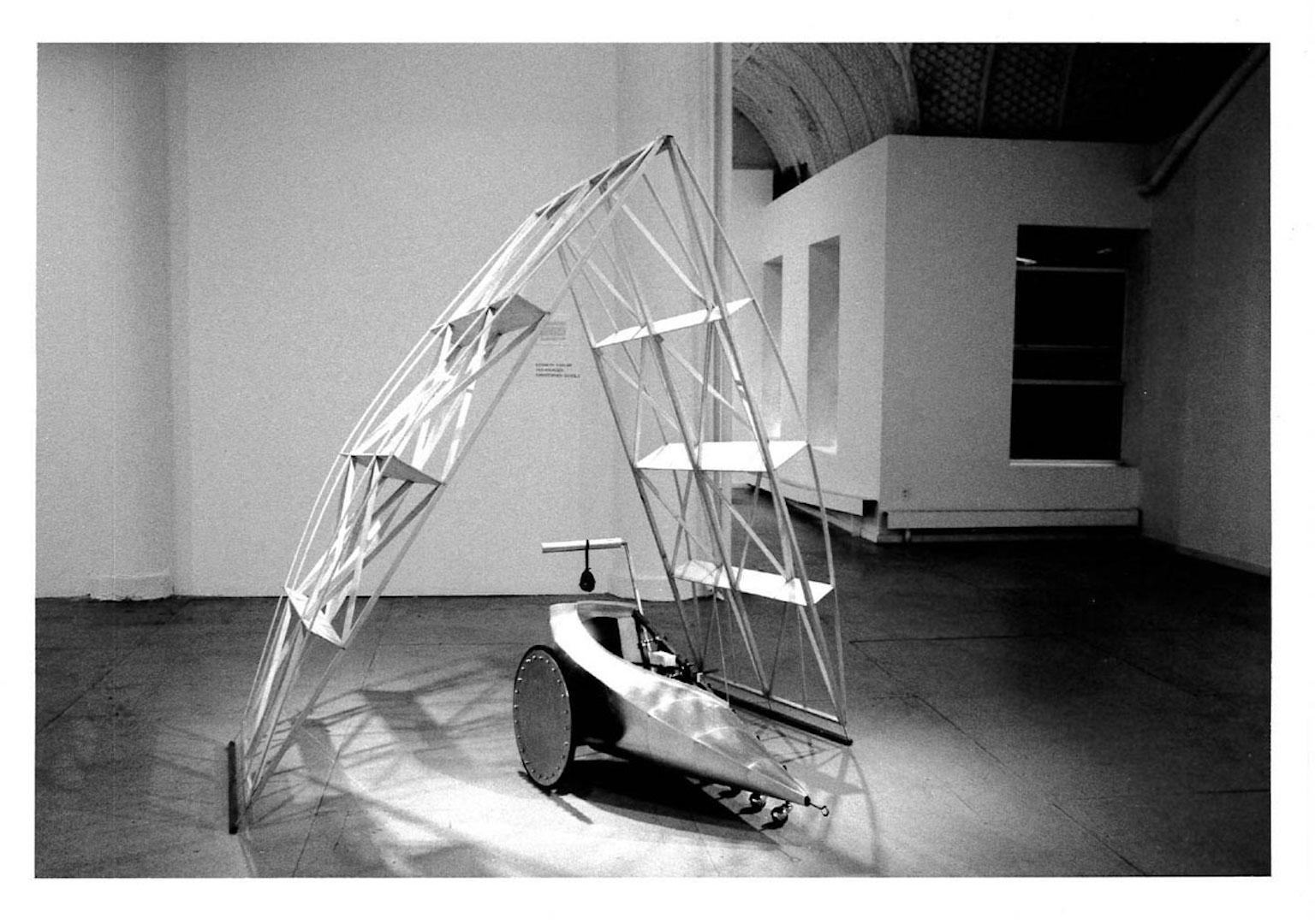 A shell of an architectural edifice stands over a small moveable two-wheeled car in a gallery space.