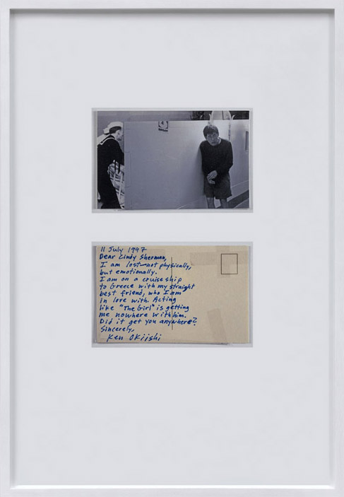 Two small items in a white glossy frame, one is a photograph and the other a postcard. The photograph is a black and white image of a man standing, slightly hunched over, pasted on top of an image of a sailor. Beneath the photograph, a faded handwritten postcard reads in blue ink, 