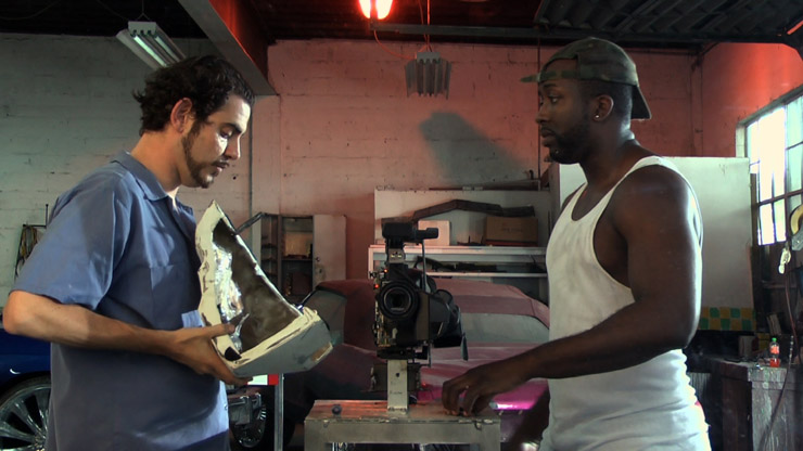 A side view of two men, facing eachother, in a workshop space. On the left, the man holds a metal object to place it onto the machine sitting inbetween them.