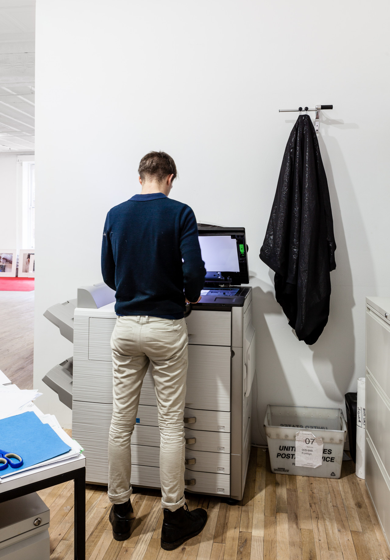 A man stands at a copy machine with the hood lifted and its light on. On a wall directly behind the copy machine, black fabric hangs from a small, horizontal pole to the right of the standing figure.