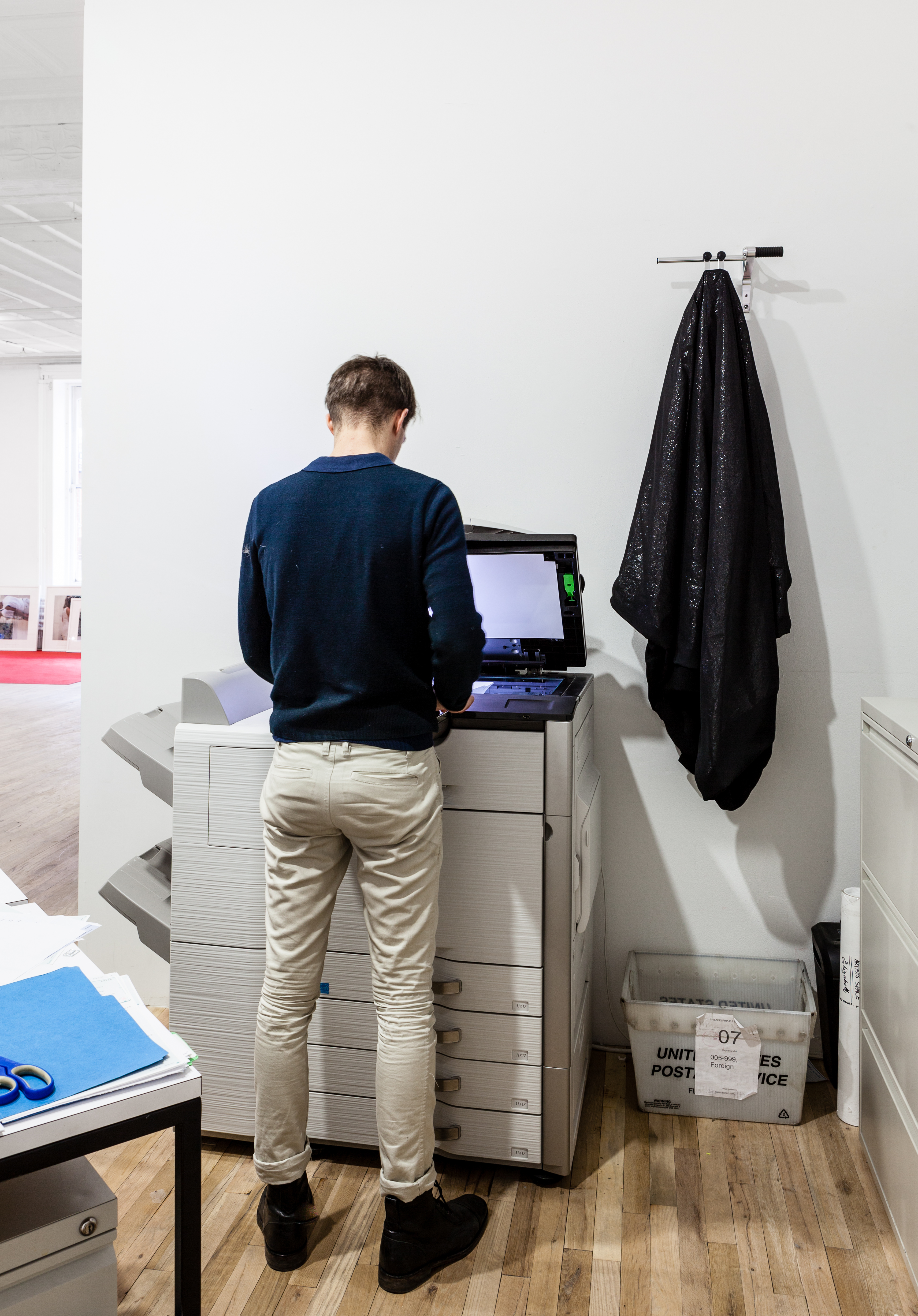 A man stands at a copy machine with the hood lifted and its light on. On a wall directly behind the copy machine, black fabric hangs from a small, horizontal pole to the right of the standing figure.