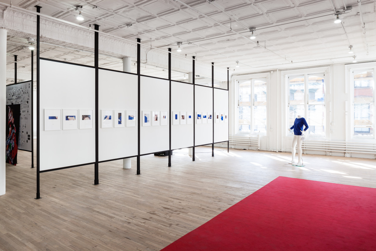 An angled view of a paneled wall displaying a long line of framed photographs in an exhibition space. A white mannequin wearing a blue shirt stands to the right of the wall. A large red carpet covers the ground in the bottom right portion of the image.