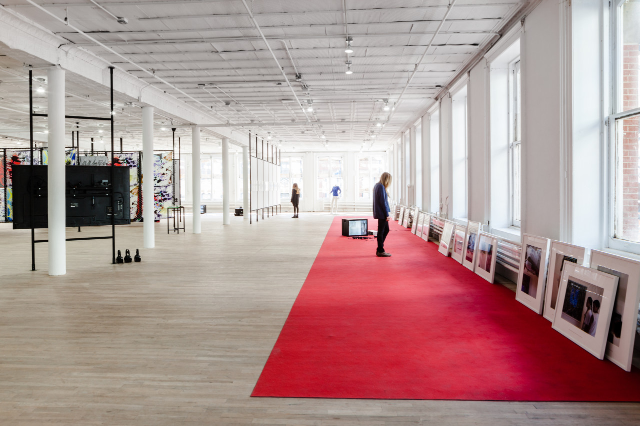 A large red carpet extends across the length of an exhibition space next to a windowed wall. A long line of framed photographs lean against the windowed wall. Two figures examine the photographs and additional artworks displayed in the space