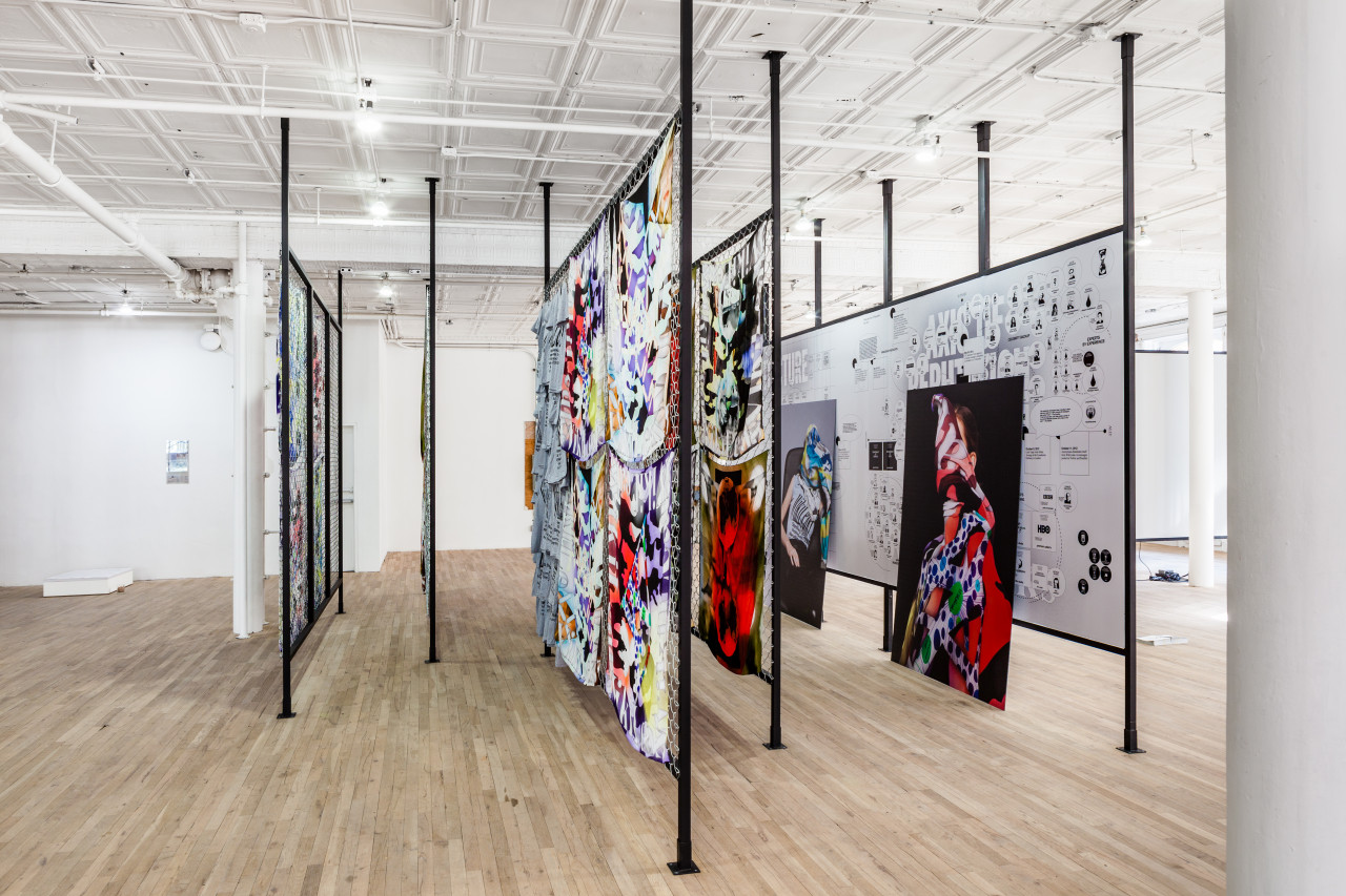 An angled view of several dividers displaying artworks in the middle of a room. Colorful textiles hang from two of the central dividers, while several framed photographs lean against rightmost divider.