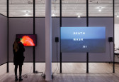 Two video monitors installed on a freestanding wall in an exhibition space. A woman stands in front of the leftmost monitor, which displays a bright, red image.  A larger monitor to the right displays a cool-toned scene of an ocean. White text superimposed over the image reads, 