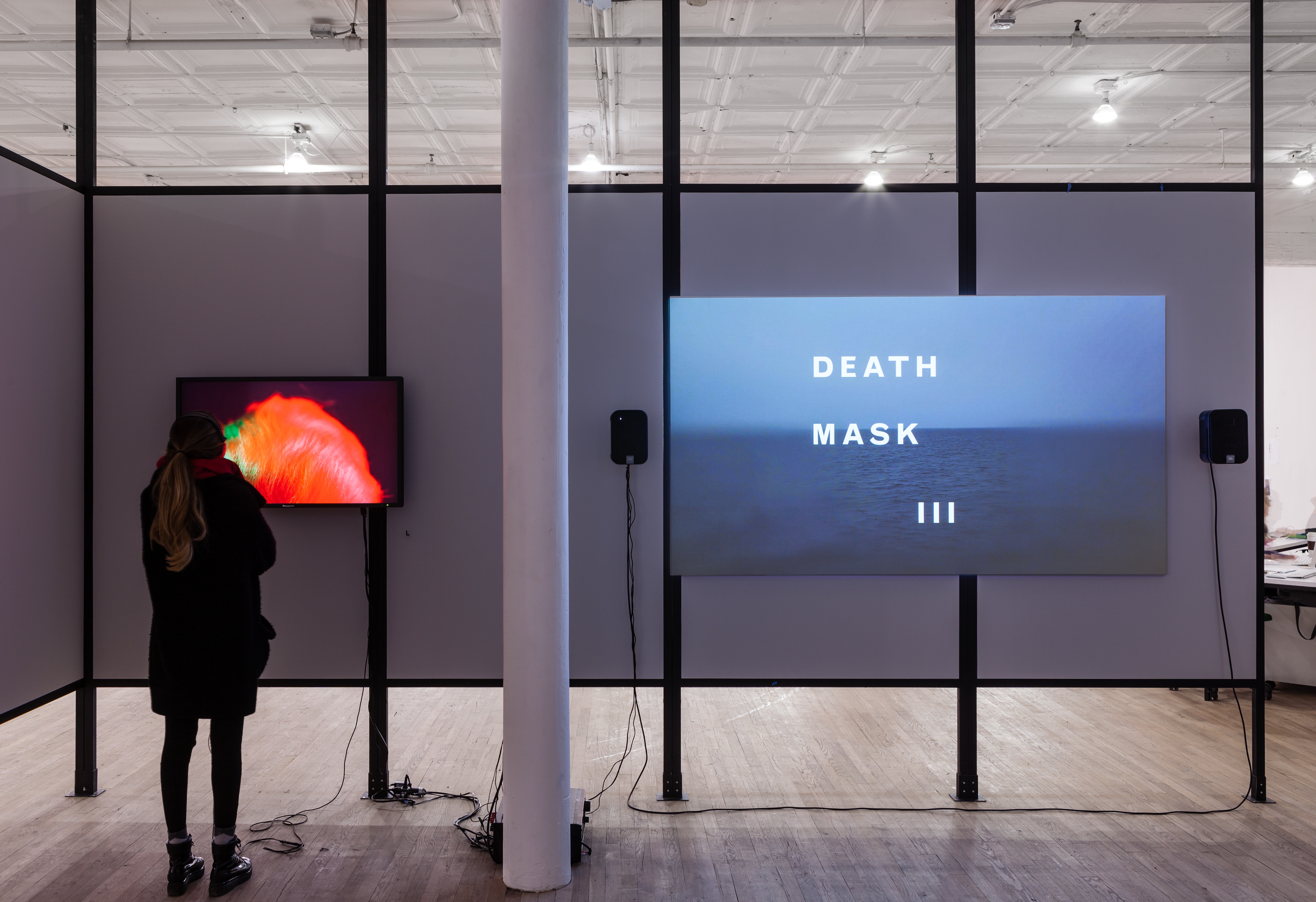 Two video monitors installed on a freestanding wall in an exhibition space. A woman stands in front of the leftmost monitor, which displays a bright, red image.  A larger monitor to the right displays a cool-toned scene of an ocean. White text superimposed over the image reads, 