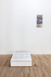 A stack of large papers filled with text sit on a low, white plank on the ground. To the right of the paper stack, a newspaper clipping is displayed on a white wall. A small glass filled with rubber bands sits on the ground to the right of the paper stack.