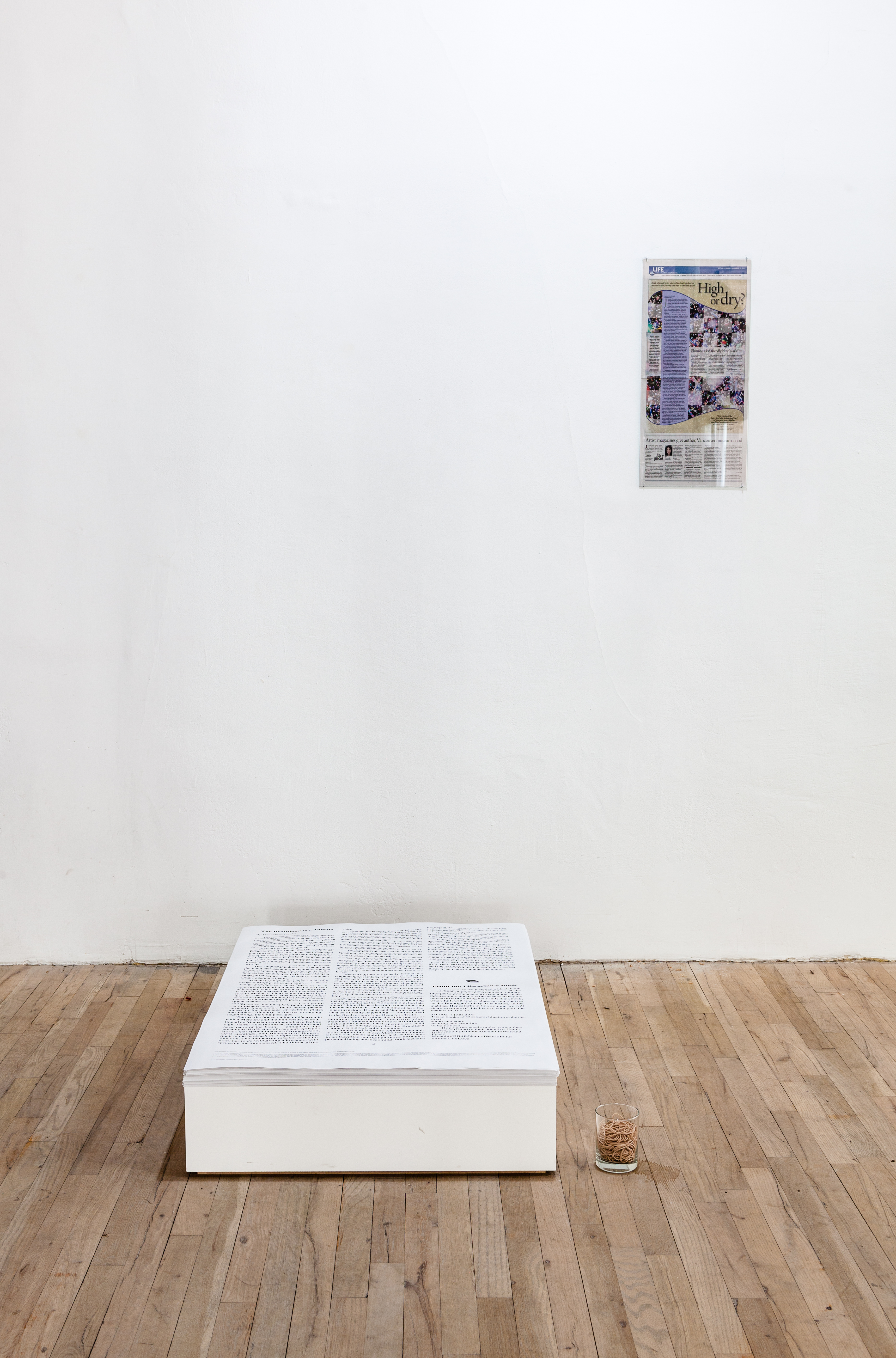 A stack of large papers filled with text sit on a low, white plank on the ground. To the right of the paper stack, a newspaper clipping is displayed on a white wall. A small glass filled with rubber bands sits on the ground to the right of the paper stack.