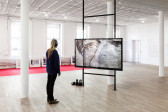 A woman wearing headphones stands in front of a horizontal monitor displaying an image of two illustrated eyes and a nose. Behind the monitor, several framed photographs lean against a wall. A long, red carpet runs across the floor underneath the photographs.