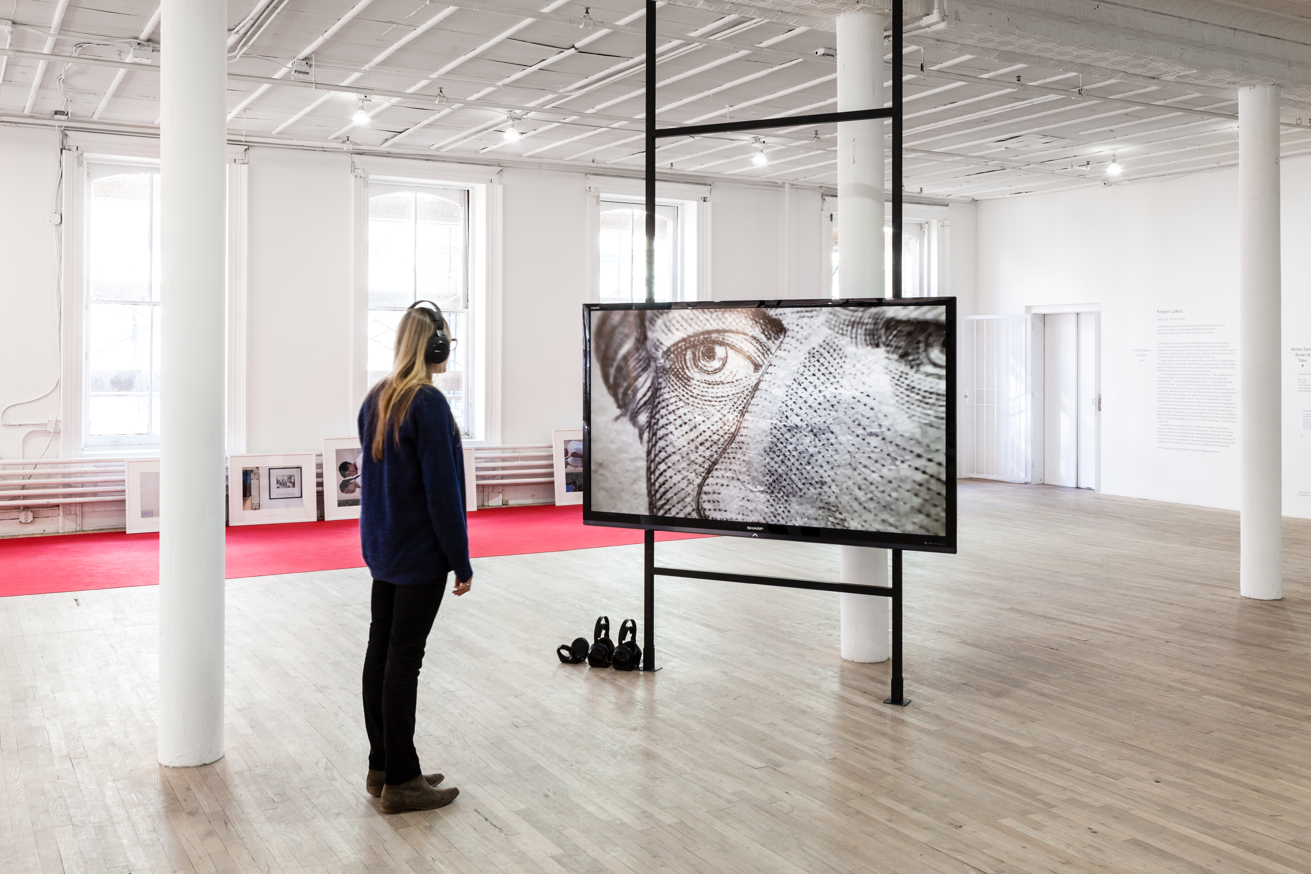 A woman wearing headphones stands in front of a horizontal monitor displaying an image of two illustrated eyes and a nose. Behind the monitor, several framed photographs lean against a wall. A long, red carpet runs across the floor underneath the photographs.