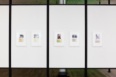 Five framed photographs and documents displayed on freestanding, white panels in an exhibition space.