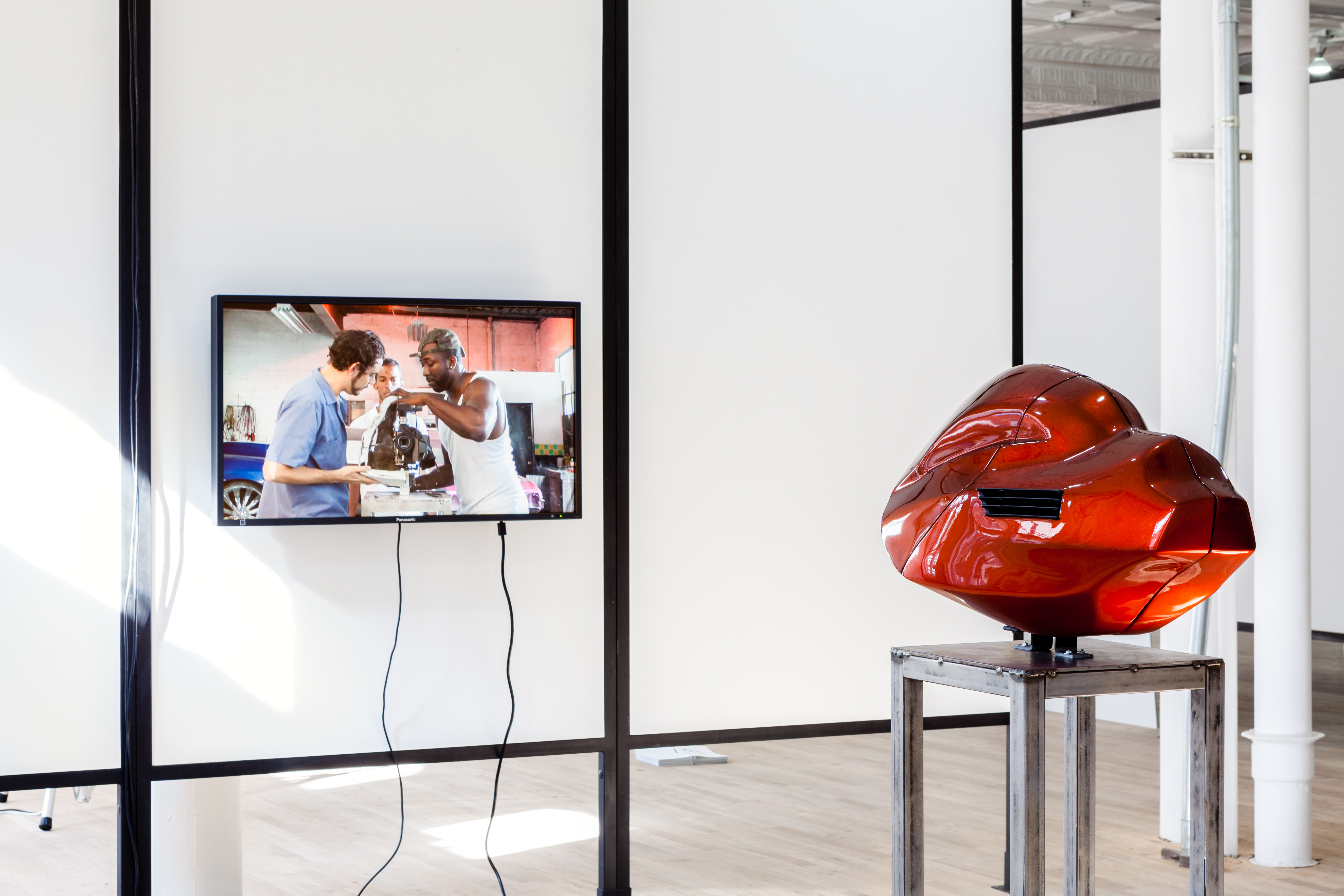 A red metal sculpture displayed on a pedestal in front of a monitor installed on a freestanding wall. The monitor's screen displays an image of three people gathered around the displayed sculpture as it is pieced together.