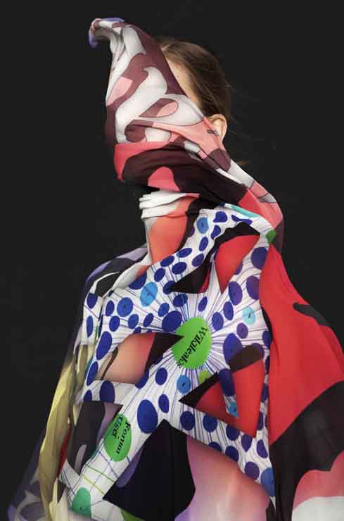 A portrait of a woman whose face is covered by a colorful cloth that extends over her entire top body. Only the top of her ear and hair are visible.