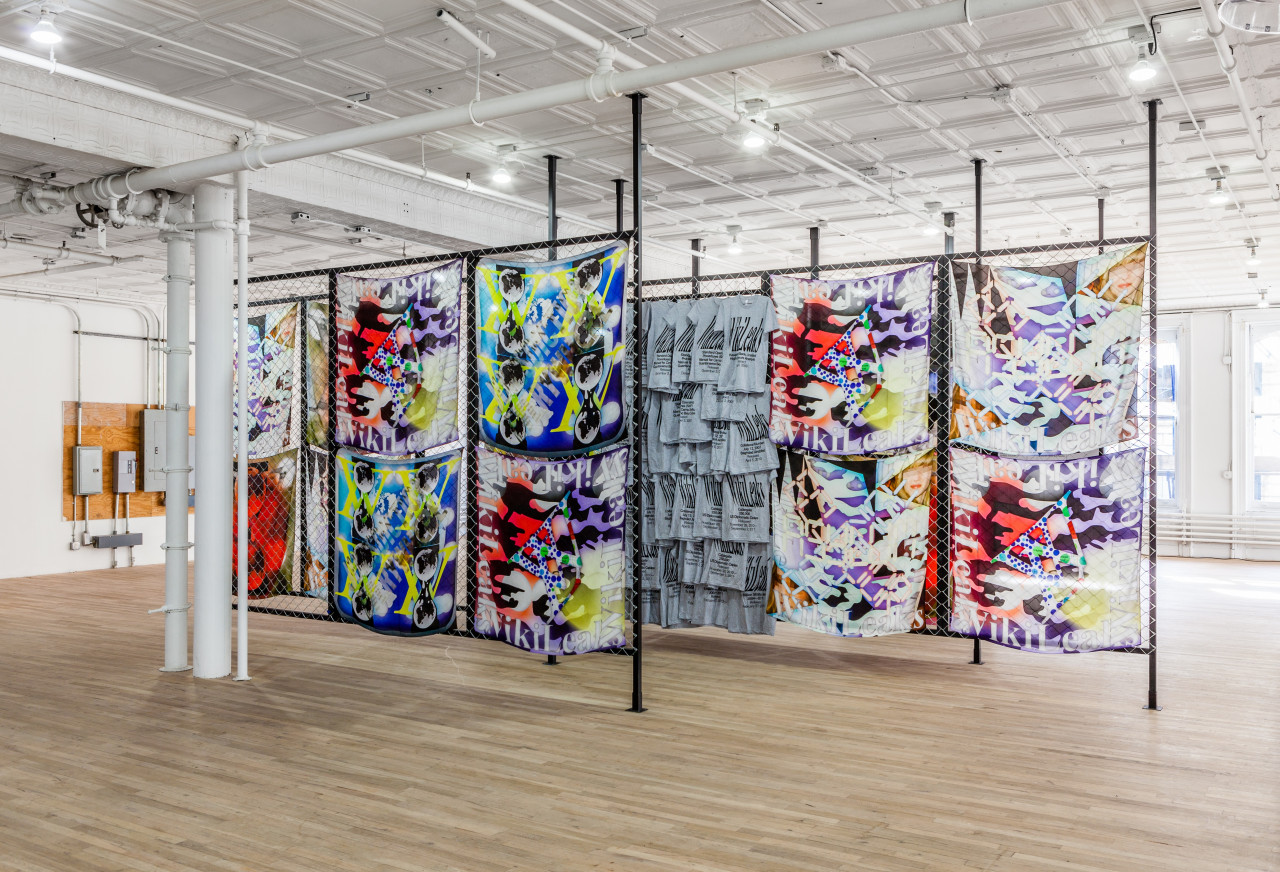 Several parallel dividers constructed from metal fencing positioned between black metal frames are arranged in an exhibition space. Colorful textiles and t-shirts are displayed on the fencing.