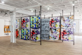 Several parallel dividers constructed from metal fencing positioned between black metal frames are arranged in an exhibition space. Colorful textiles and t-shirts are displayed on the fencing.