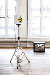 A white speaker displayed on a three-legged stand in an exhibition space. To the right of the stand, a television monitor rests on the ground. An image of gravel and dirt is pictured on its screen.