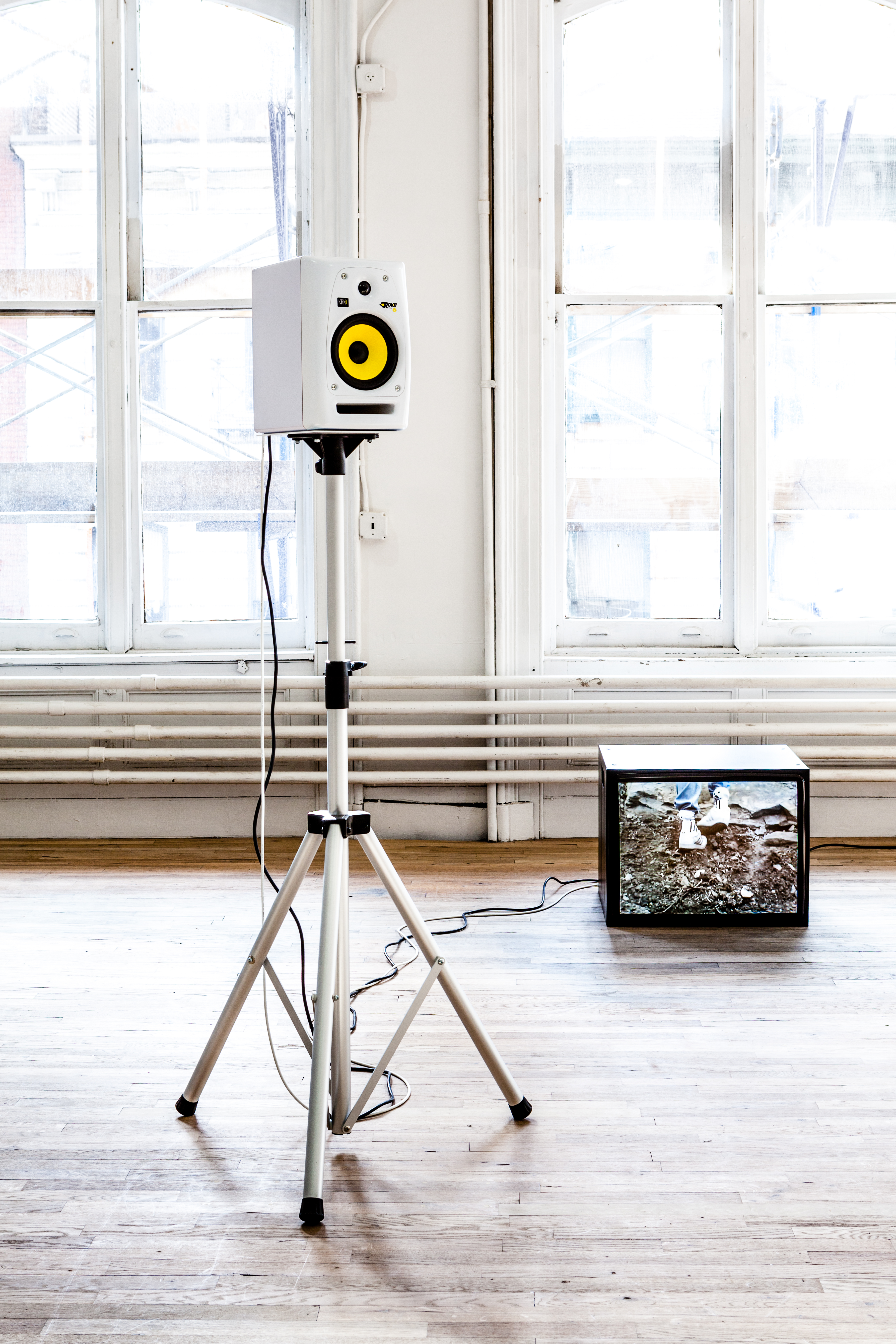 A white speaker displayed on a three-legged stand in an exhibition space. To the right of the stand, a television monitor rests on the ground. An image of gravel and dirt is pictured on its screen.