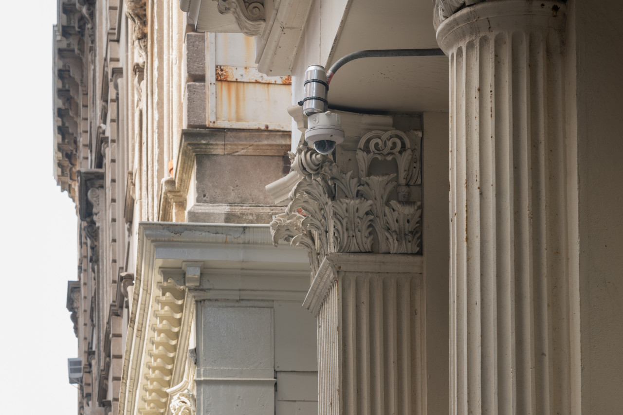 The exterior corner of Artists Space shows a building with highly elaborated columns. On the White Street side, an aluminum can is strapped to the building’s security camera.