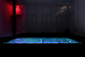 In a large dark room, a modular structure is recessed into a raised floor covered with black carpet. The structure is lit in bright blue light, and hosts open water. Behind, a projected image from a camera obscura displays the alley outside. Red light floods the back corner of the room.