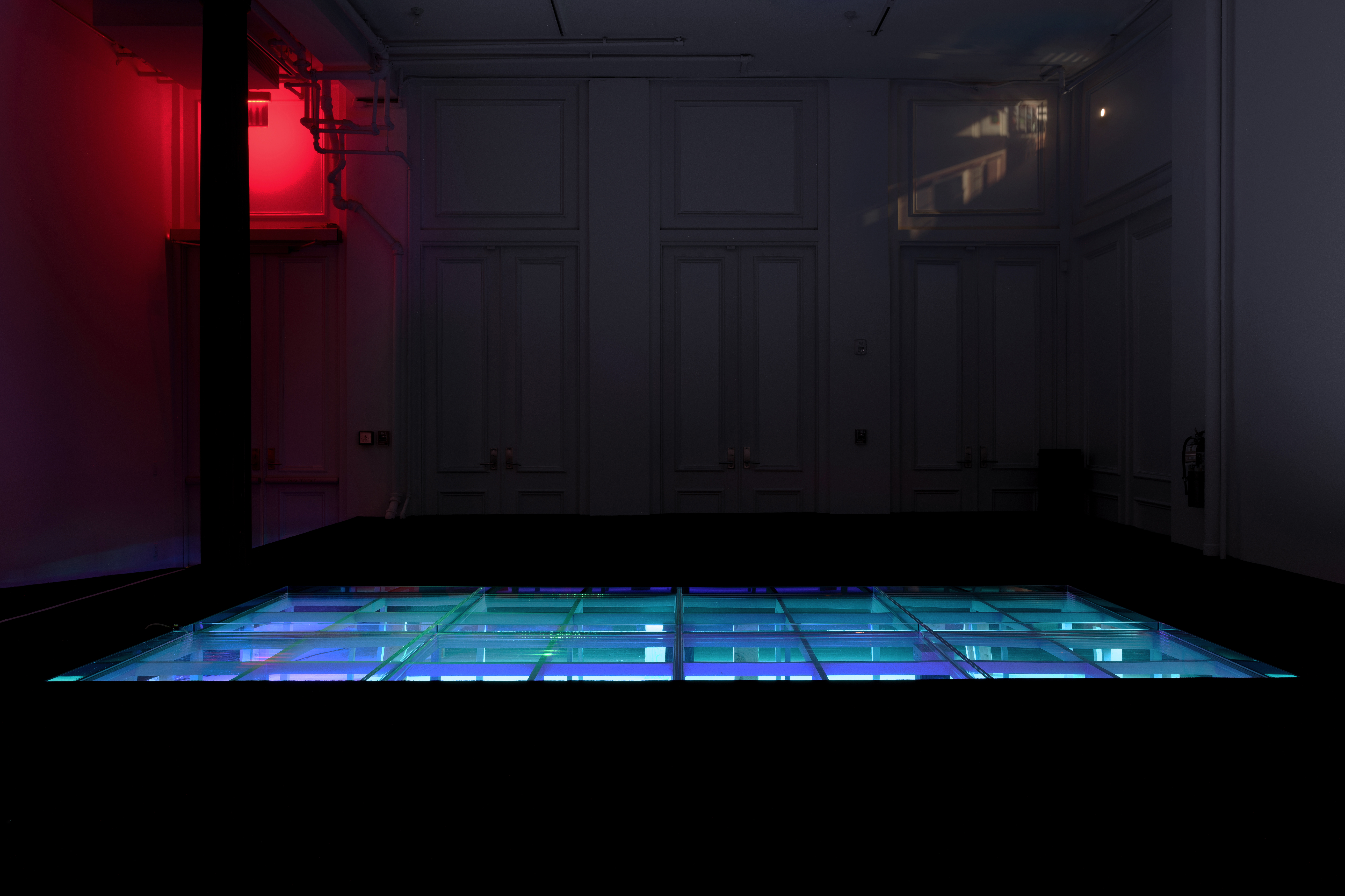 In a large dark room, a modular structure is recessed into a raised floor covered with black carpet. The structure is lit in bright blue light, and hosts open water. Behind, a projected image from a camera obscura displays the alley outside. Red light floods the back corner of the room.
