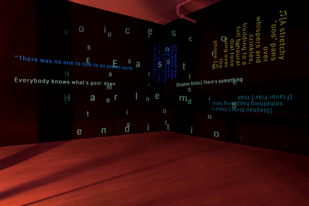 In a darkened room lit softly by red light, overlapping lines of open captions are projected on two adjacent black walls. The text varies in color (in faint greens, yellows and blues) and size.