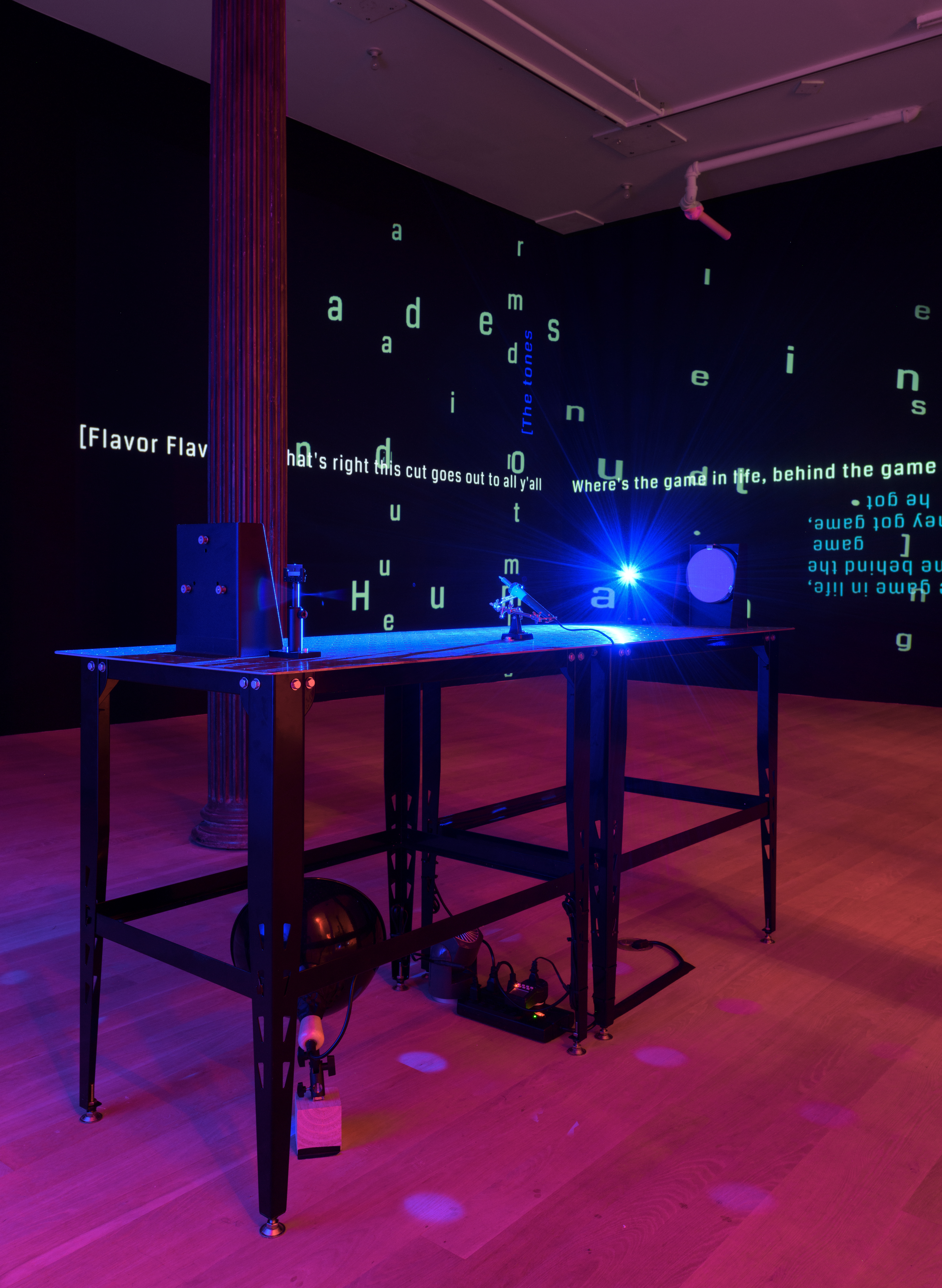 In a darkened room, a bright blue LED light, two circular mirrors and scientific mounts sit atop two metal welding tables. Behind, on two adjacent black walls, lines of projected open captions overlap in varying directions.