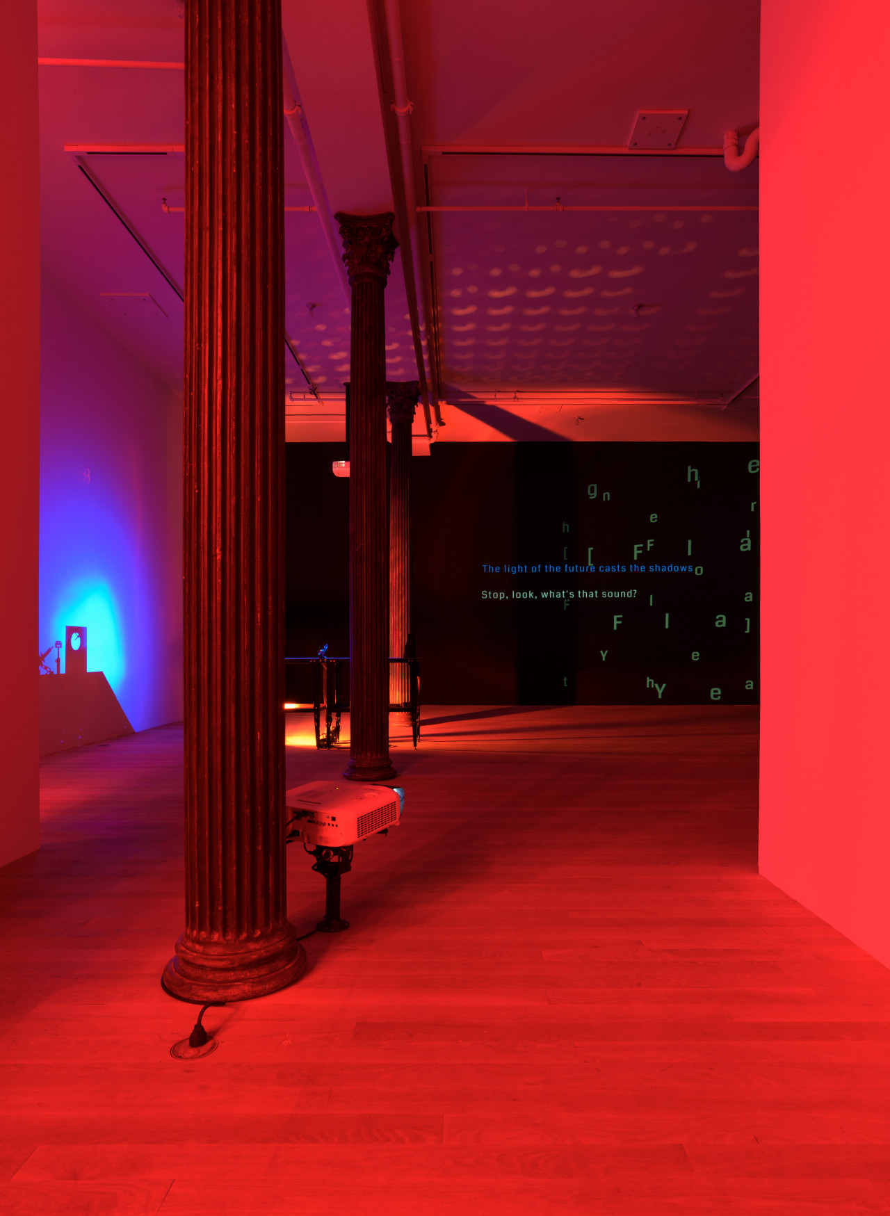 An installation view shows a darkened space flooded in red light. Between two ornate columns, a projector is mounted close to the ground on a short stand. An iridescent blue glows around a shadow cast by a scientific experiment on the left wall behind, while the ceiling is covered in warm crescent silhouettes. On the back black wall, lines of projected text overlap in varying directions.