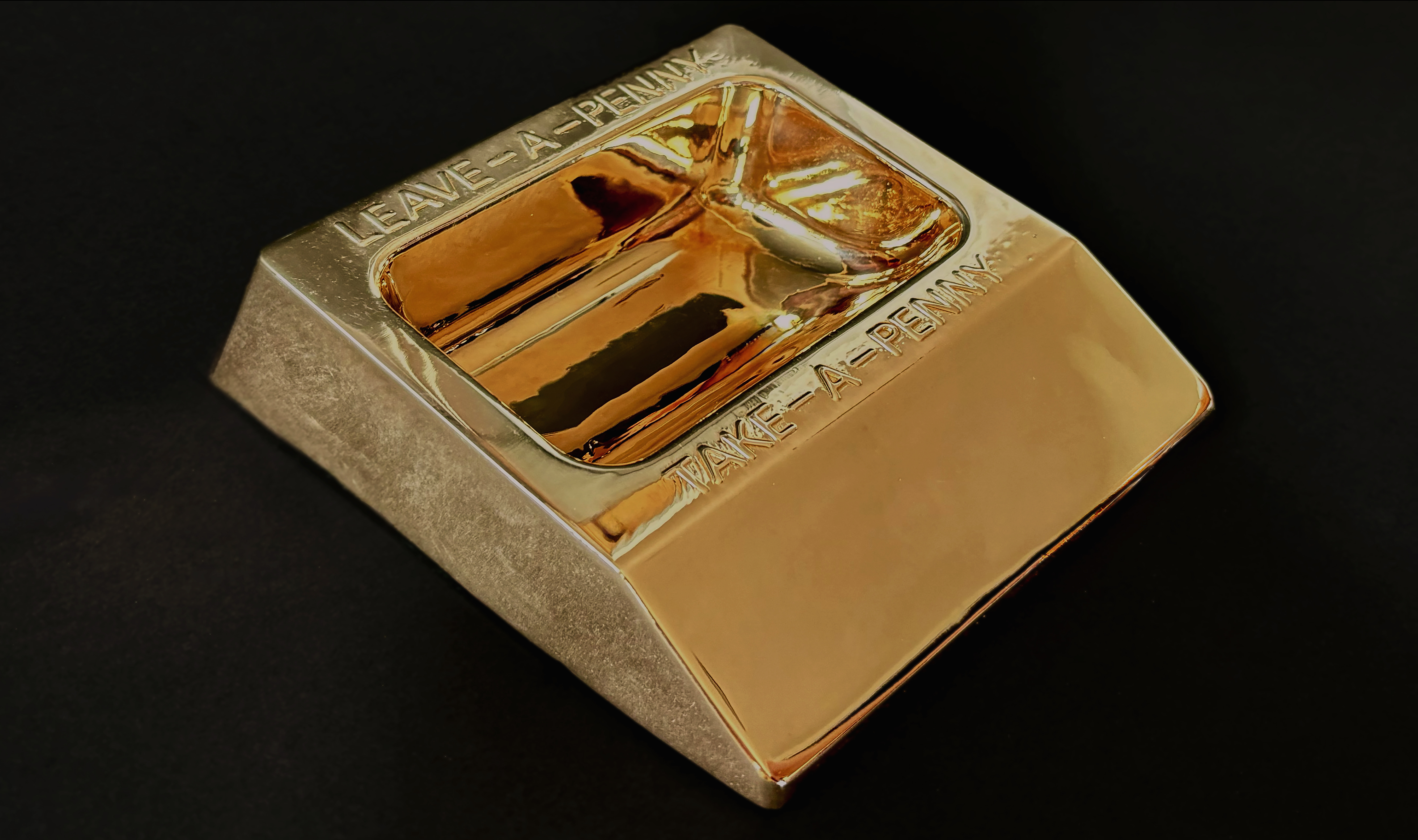 A common convenience store countertop coin share dish shines in solid bronze on a soft black velvet ledge. Its raised text reads: “LEAVE - A - PENNY / TAKE - A - PENNY”.