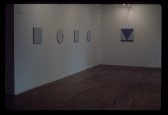 A color installation image of a gallery space. Several small canvases painted with geometric forms are hung on the walls.