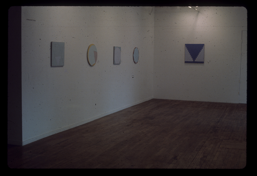 A color installation image of a gallery space. Several small canvases painted with geometric forms are hung on the walls.
