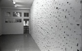 A photograph of small metal circles, bars, and other pieces mounted on a white gallery wall.