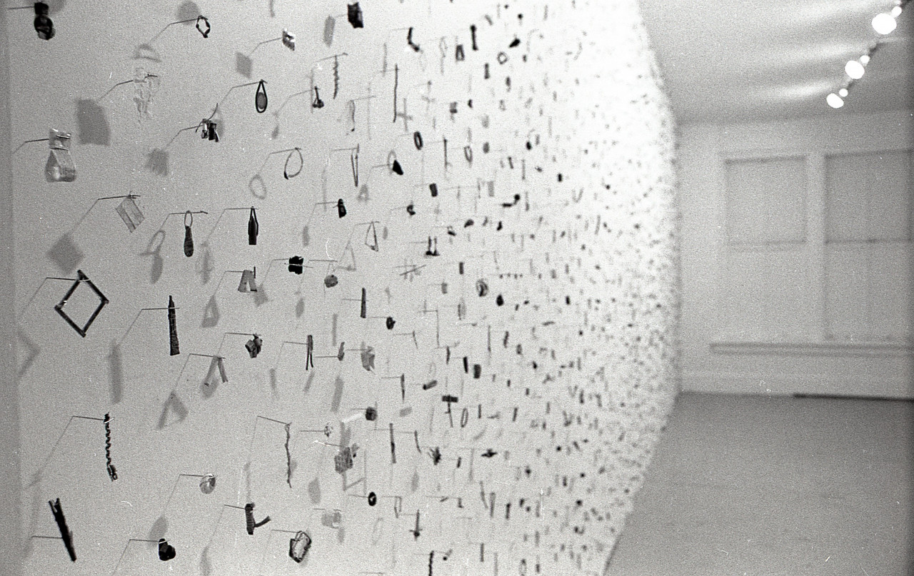 A photograph of small metal circles, bars, and other pieces mounted on a white gallery wall.