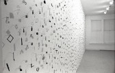 A photograph of small metal circles, bars, and other pieces mounted on a white gallery wall.