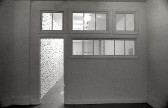 A photograph of a room painted white with an open door. In the adjacent room, small metal circles, bars, and other pieces mounted on a white gallery wall can be seen.