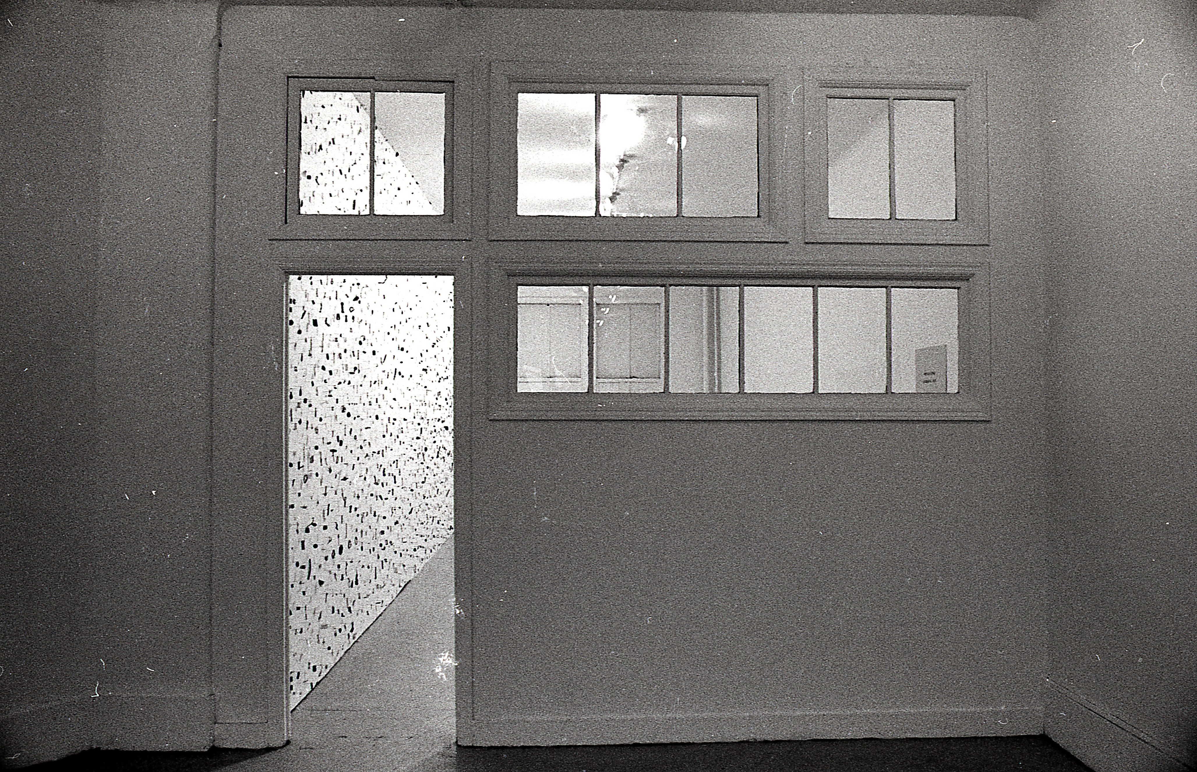 A photograph of a room painted white with an open door. In the adjacent room, small metal circles, bars, and other pieces mounted on a white gallery wall can be seen.