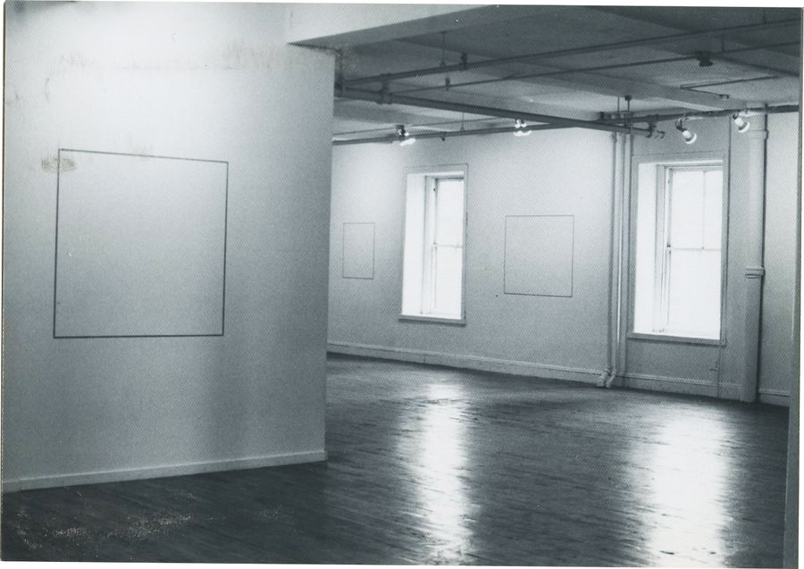 A black and white installation photograph of a gallery space. A square blank canvas hangs from the wall on the left, and two windows are visible on the right.