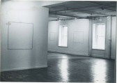 A black and white installation photograph of a gallery space. A square blank canvas hangs from the wall on the left, and two windows are visible on the right.