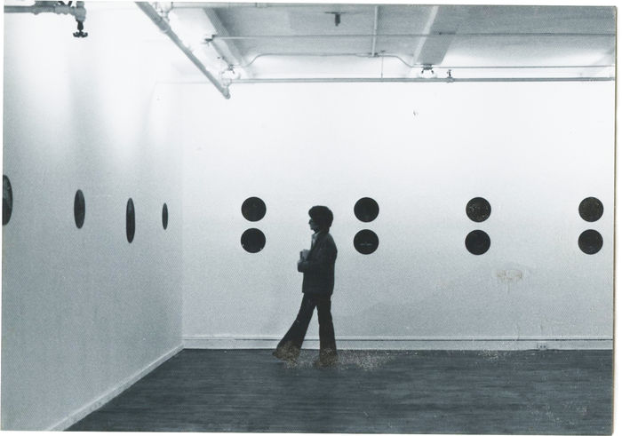 Black and white installation photograph of a gallery space. Black circles are attached to the wall and a figure is visible standing in the space.