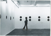 Black and white installation photograph of a gallery space. Black circles are attached to the wall and a figure is visible standing in the space.