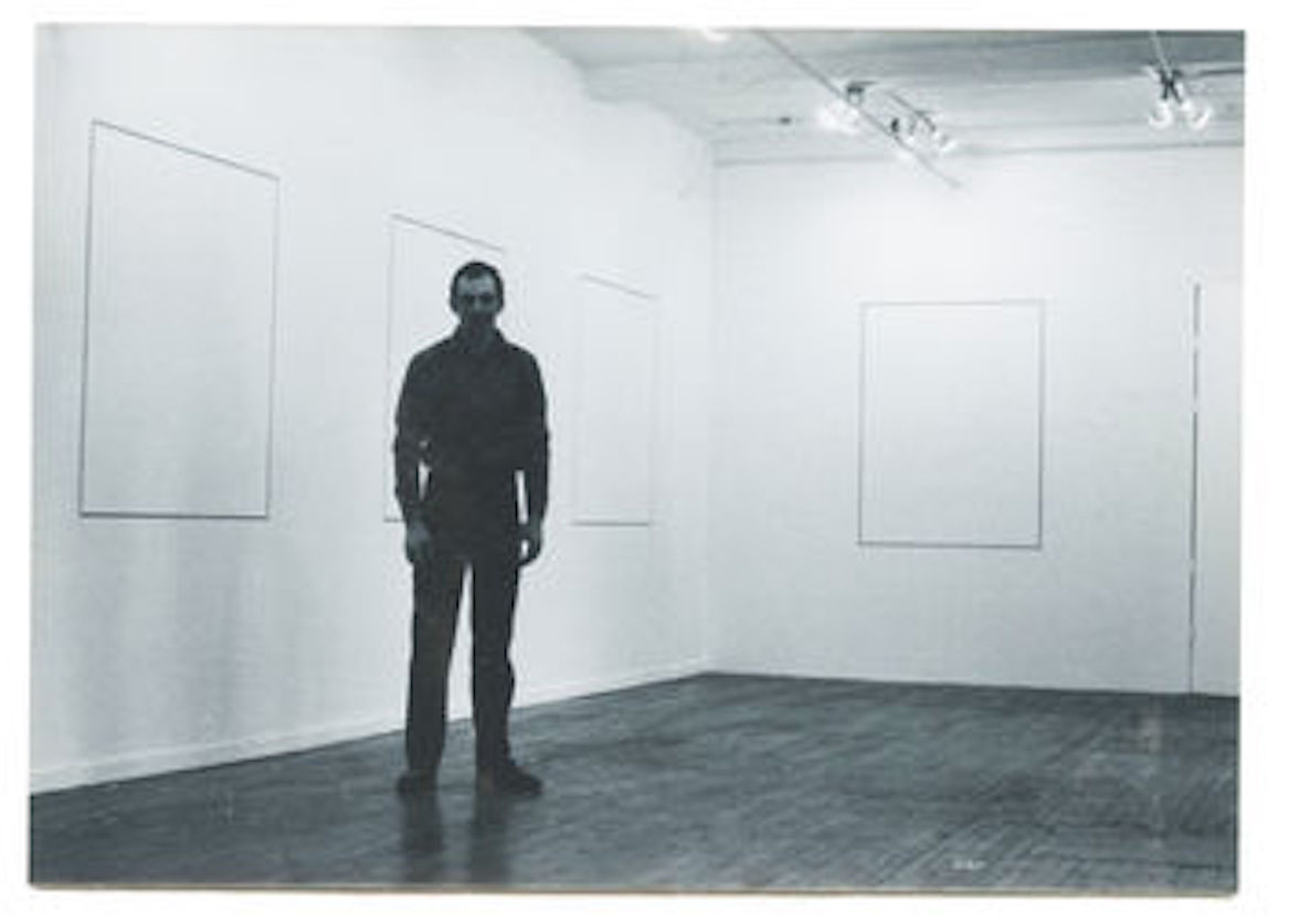 A black and white photograph of a figure standing in a gallery space, looking towards the camera. On the wall behind them, several canvases with no discernable markings hang.