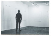 A black and white photograph of a figure standing in a gallery space, looking towards the camera. On the wall behind them, several canvases with no discernable markings hang.