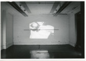 An image is projected onto a white wall. In the image, a figure's chin and upper chest are visible as they hold a round object in their hand, close to their chin.