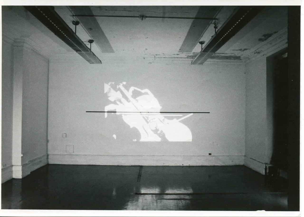 An image is projected onto a white wall. In the image, a figure plays a cello.