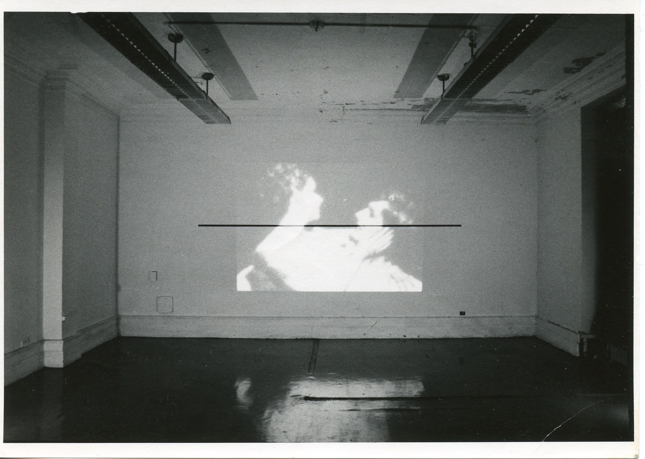 An image is projected onto a white wall. In the image, a woman holds a man's head in her hands.