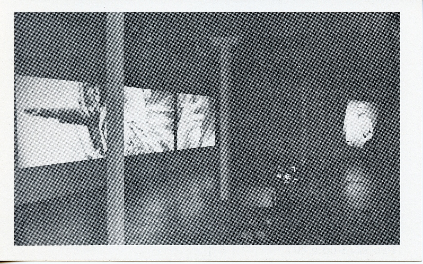 A black-and-white photograph of a video installation with multiple projections running simultaneously on different parts of the walls. A chair and table are visible in the foreground.