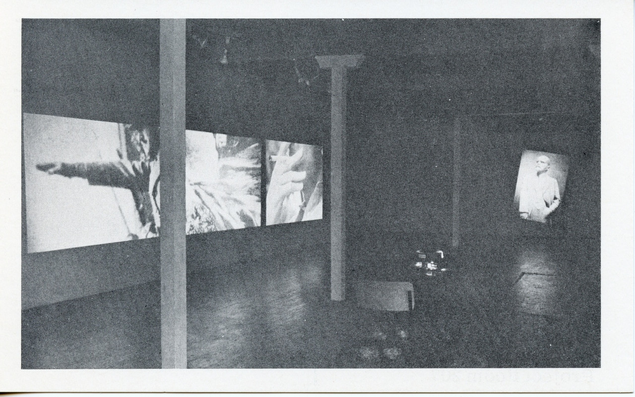 A black-and-white photograph of a video installation with multiple projections running simultaneously on different parts of the walls. A chair and table are visible in the foreground.