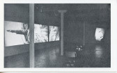 A black-and-white photograph of a video installation with multiple projections running simultaneously on different parts of the walls. A chair and table are visible in the foreground.