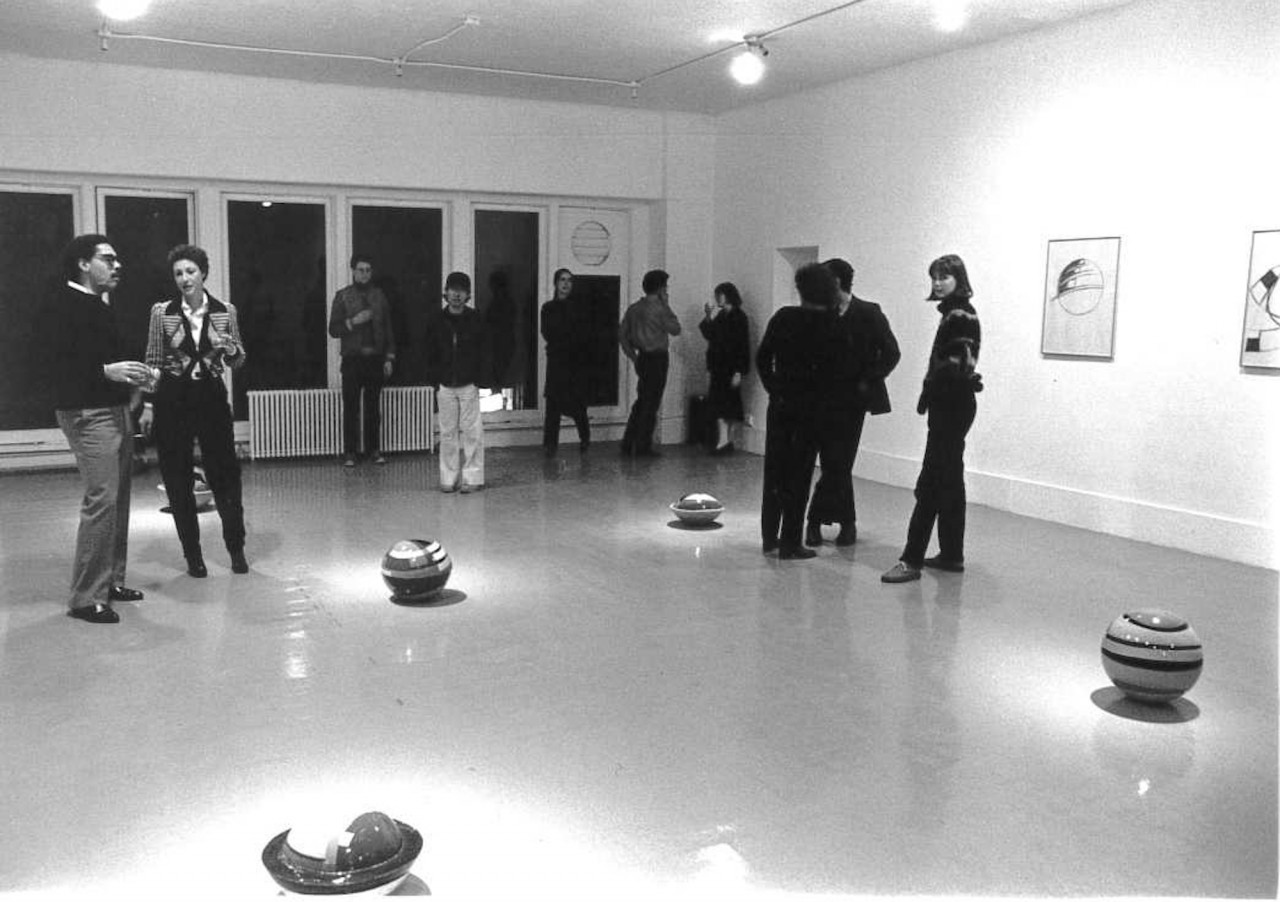 Black and white photograph of people standing in a gallery among five spherical balls on the floor. Framed artworks hang on the right wall.