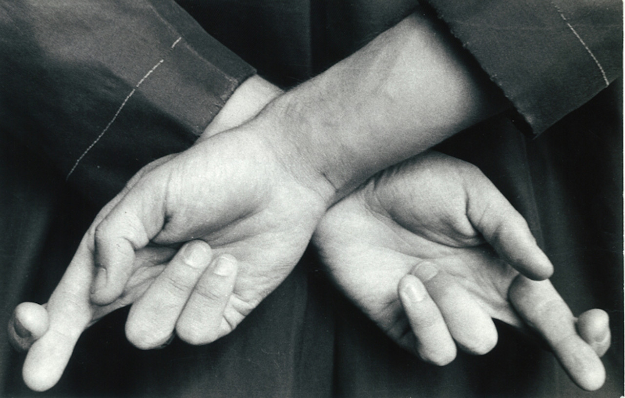 A black and white image of two hands behind a figure's back. They cross their index and middle finger on each hand.