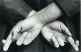 A black and white image of two hands behind a figure's back. They cross their index and middle finger on each hand.